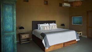 Un dormitorio con una cama grande, dos mesitas de noche, un buró, un aire acondicionado instalado en la pared, una lámpara colgante y un cuadro enmarcado.