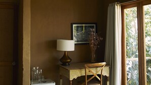 Una habitación con una lámpara colgante de mimbre, un escritorio con lámpara, una silla, un pequeño armario blanco y una ventana con cortinas.