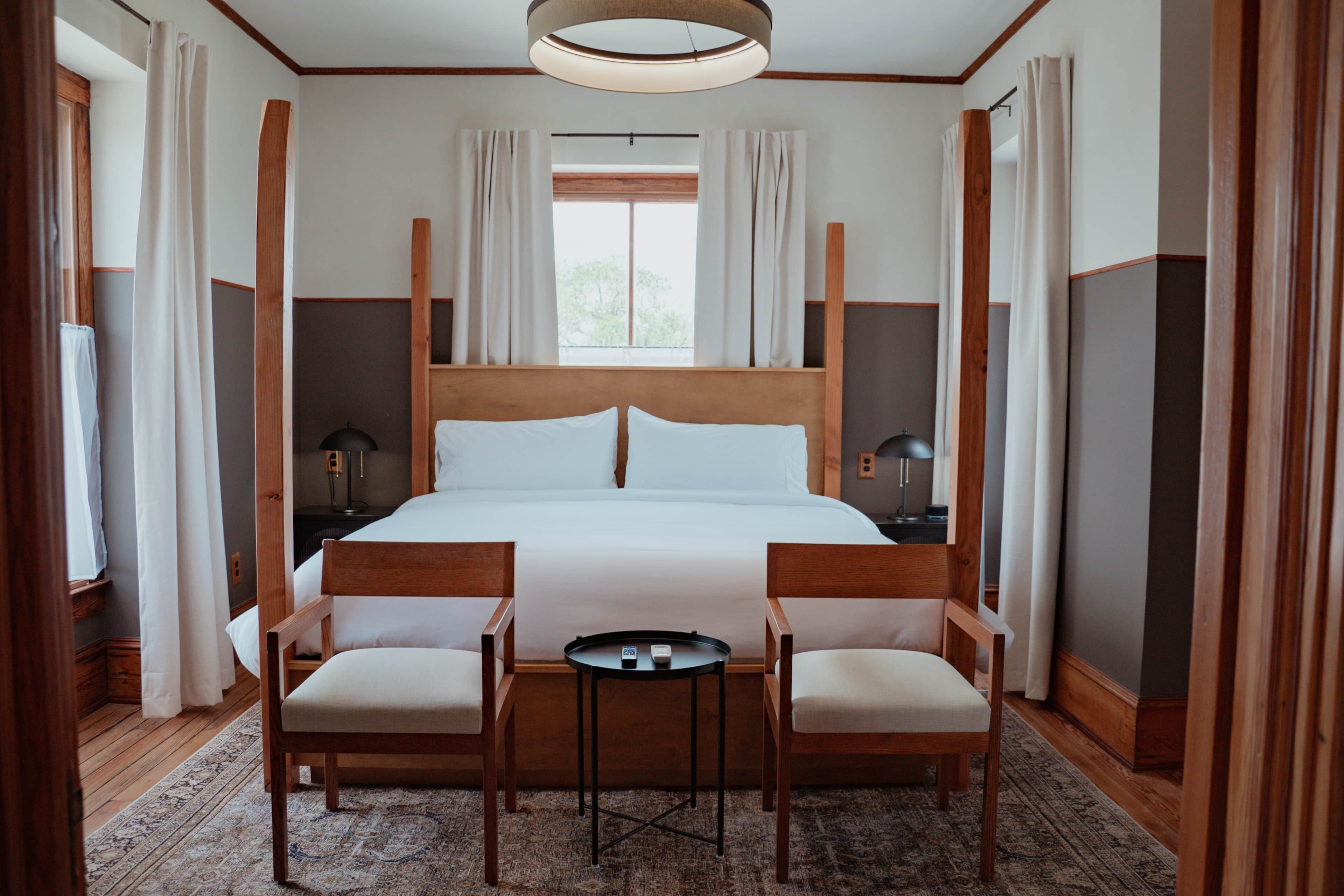 Una habitación de hotel con una cama grande, dos sillas de madera, una mesita pequeña y una mesita de noche con lámpara.