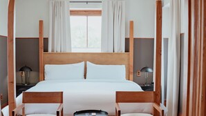 Ein Hotelzimmer mit einem großen Bett, zwei Holzstühlen, einem kleinen Tisch, einer Deckenleuchte und einem fenster mit Vorhängen.