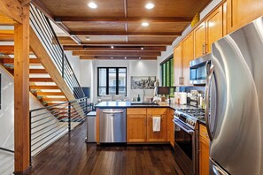 A modern kitchen with wooden cabinets, stainless steel appliances, a central island, and a staircase with a railing. - Green Lake Modern K/Q  + Parking | Stadium Access (Seattle)