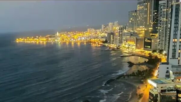 Paisagem urbana costeira ao anoitecer, com edifícios iluminados e uma praia.