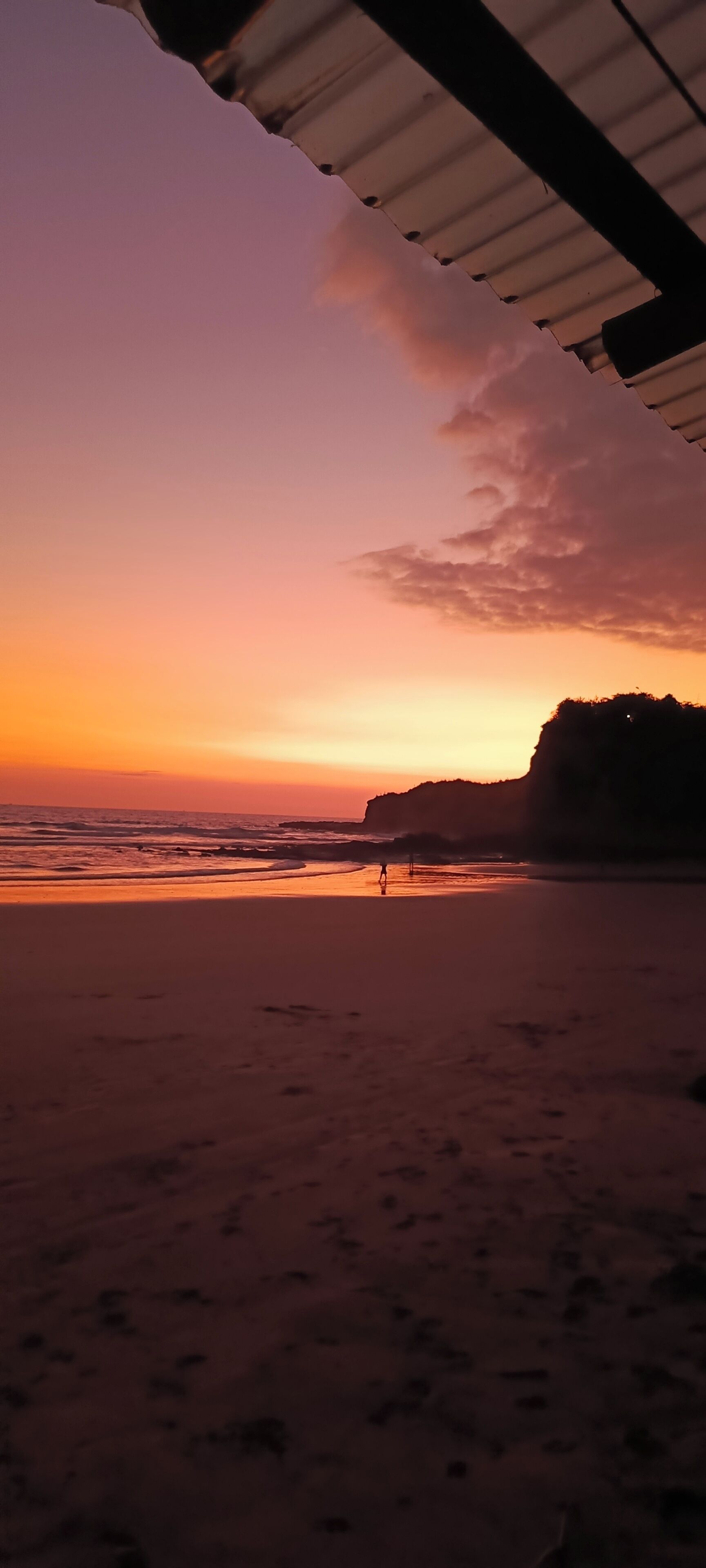Escena de playa con atardecer, una persona caminando y una estructura con techo.