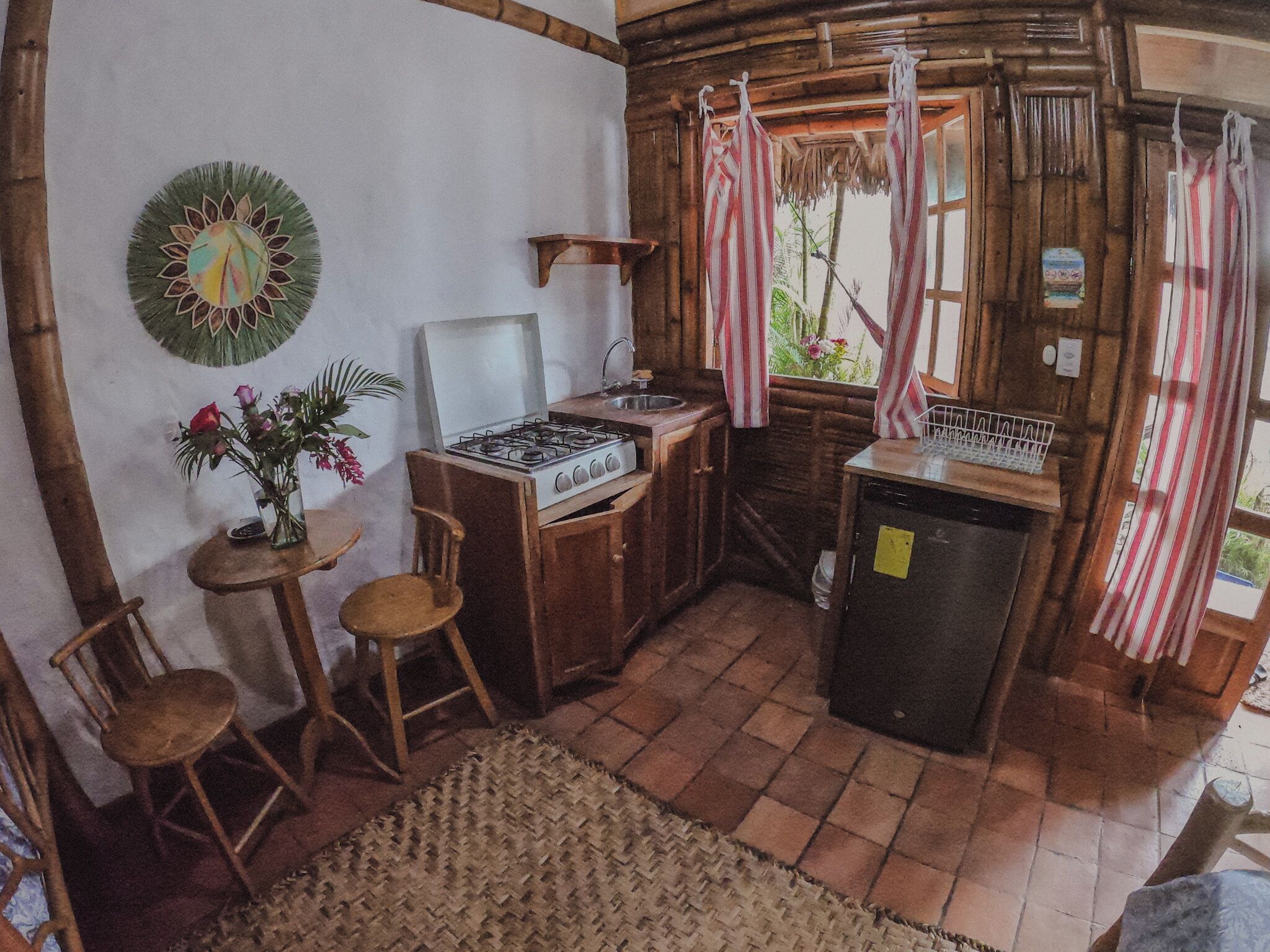 A rustic kitchen with a gas stove, wooden chairs, a small table with a vase, and a window with curtains.