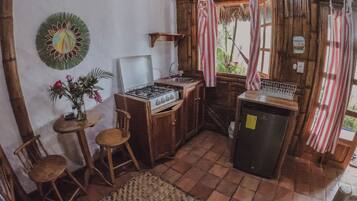 A rustic kitchen with a gas stove, wooden chairs, a small table with a vase, and a window with curtains.