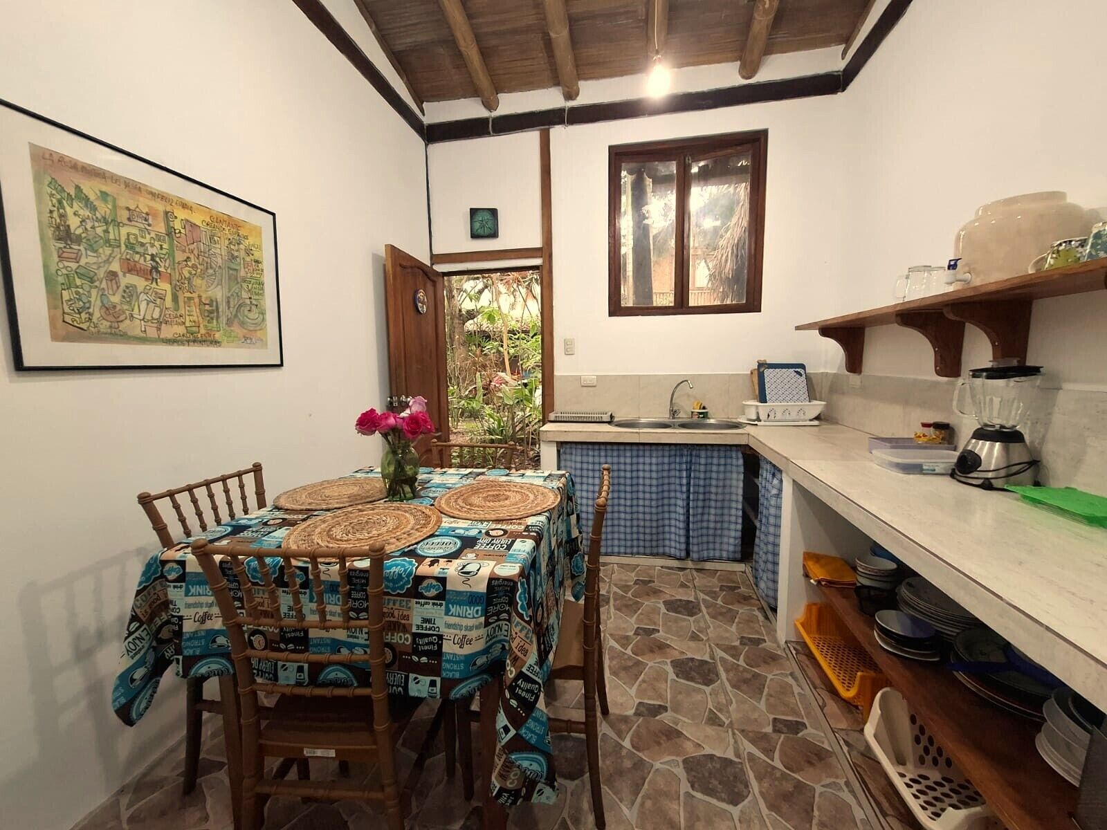 A kitchen with a stone floor, a dining table with a coffee-themed tablecloth, and a window with a view of greenery.