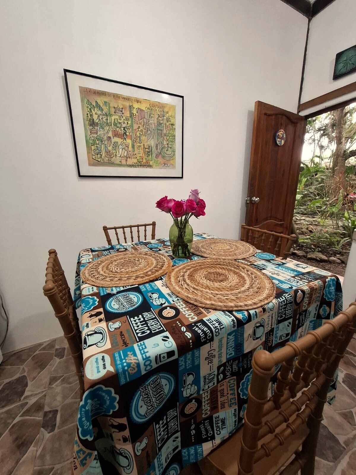 Une table à manger recouverte d’une nappe aux motifs de café, une œuvre d’art encadrée au-dessus, et une porte en bois sur la droite.