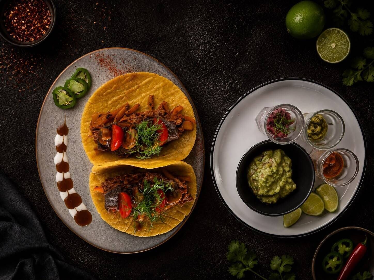 Two tacos on a plate with lime wedges and a bowl of guacamole.