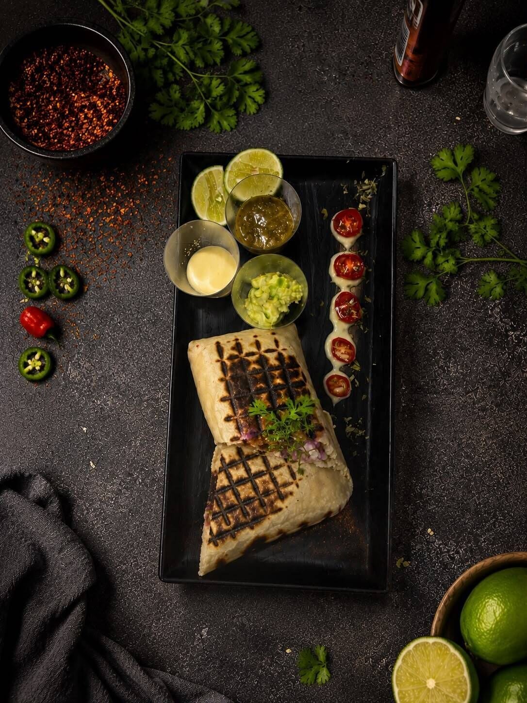Un burrito garni de divers ingrédients et accompagné de sauces, présenté sur une assiette sombre.