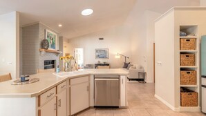 A modern kitchen with a built-in dishwasher, a white brick accent wall, and a dining area with a sofa and table. - New Reno! Landing Pad-4 lrg BR-private heated pool-FlyIn-Golf-Daytona Speedway (Port Orange)