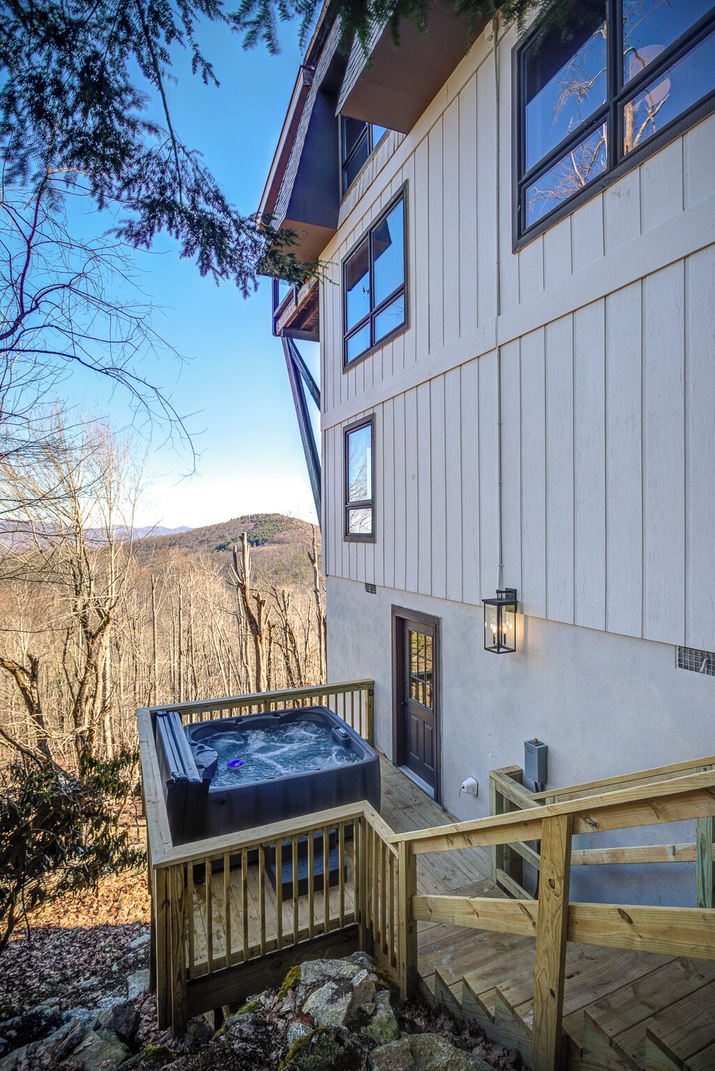 A two-story house with a hot tub on the deck, a wooden staircase, and a view of the surrounding forest and hills.