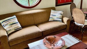 A living room with a brown sofa, two pillows, a round window, a red door, a wicker chair, and a wooden coffee table. - Riverside oasis with private pool and backyard.  Walking distance to the beach. (Paquera)
