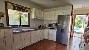 A kitchen with white cabinets, a stainless steel refrigerator, a microwave, and a window with blinds. - Riverside oasis with private pool and backyard.  Walking distance to the beach. (Paquera)