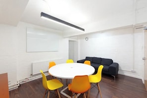 Ein Konferenzraum mit einem weißen Tisch, orangefarbenen Stühlen, einer schwarzen Couch und einer Tafel. Weiße Backsteinwände.