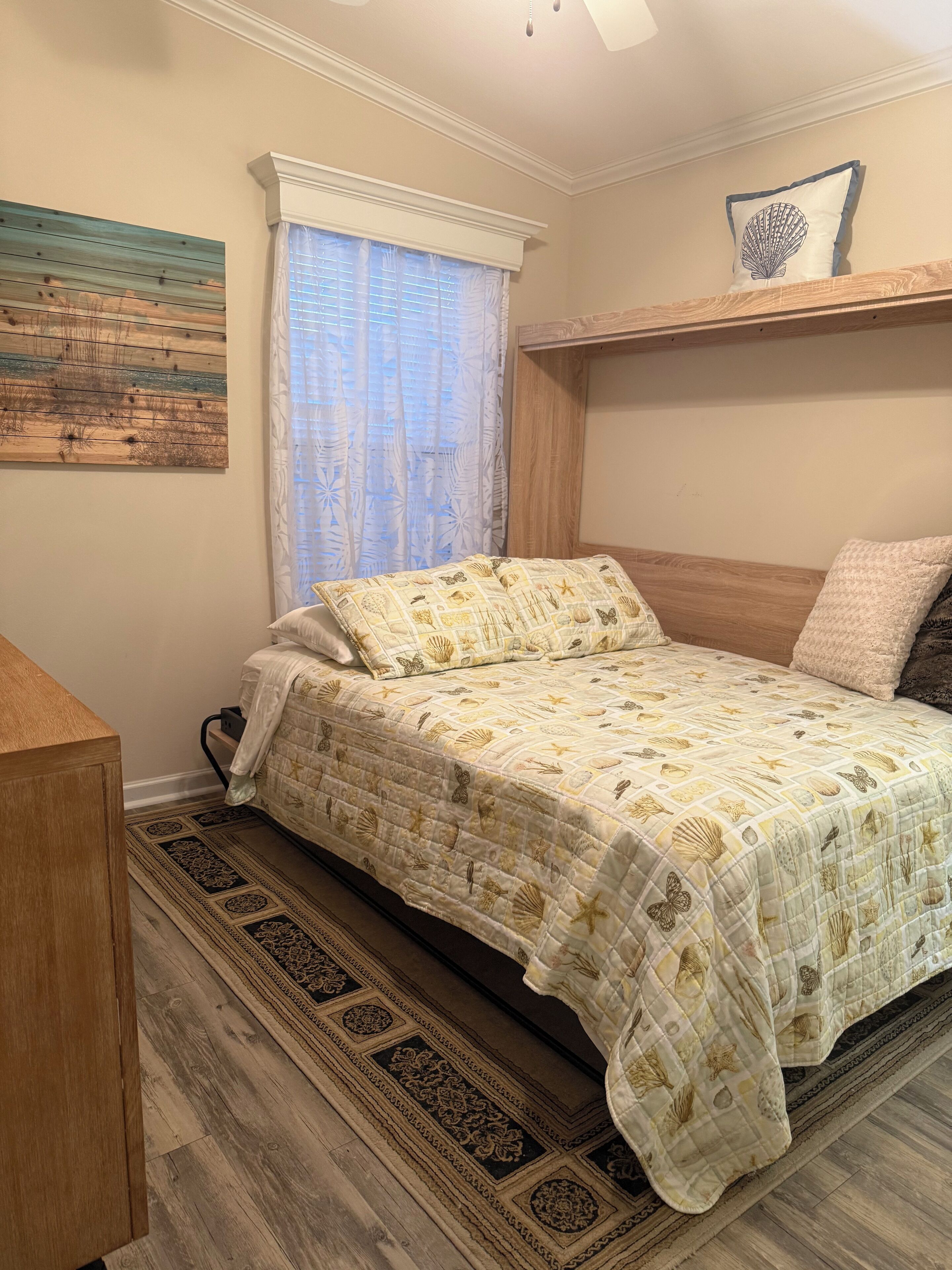 A bedroom with a bed, a wooden headboard, a window with sheer curtains, a wall-mounted shelf with a decorative pillow, and a patterned rug.