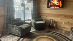 Ein gemütliches Zimmer mit Holzwänden, einem Kamin, zwei Sesseln, einer Stehlampe und einem Teppich.