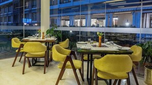 Een moderne eetruimte met gele stoelen en tafels, grote ramen en uitzicht op een gebouw met balkons.