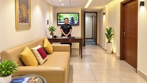 Un vestíbulo de hotel con una recepción, un sofá con cojines, una pantalla de televisión y plantas en macetas.