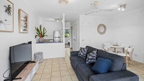 A modern living room with a flat-screen TV, a grey sofa with patterned cushions, a dining area with a white table and chairs, and a ceiling fan. - Coconut Vibes- Overlooking Resort Pool Ocean Glimpses, Holloways Beach - Cairns (Holloways Beach)