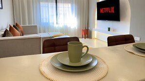Uma sala de estar moderna com um sofá, uma mesa de centro com uma caneca, uma TV exibindo 'NETFLIX' e uma área de jantar com um jogo americano.