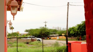 A view from a porch with a red wall, a concrete floor, and a metal fence. There is a cactus in a pot and a street visible in the background. - LIMITED OFFER FOR FIRST GUESTS - ENJOY MY HOME AT A GREAT RATE! (Noord,)