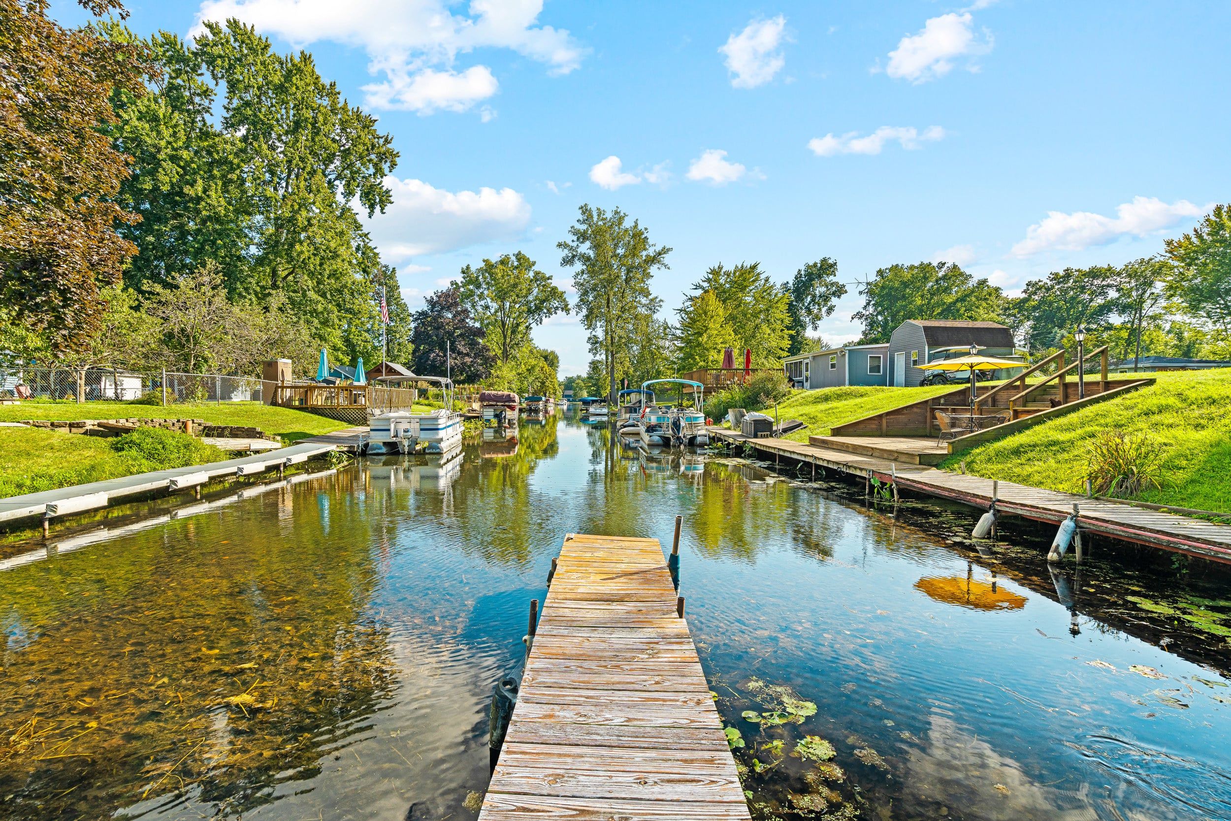 A canal with boats docked on the right, a wooden dock in the foreground, and buildings on the left.