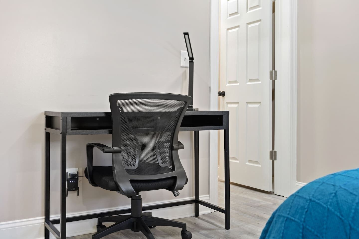 A black office chair with a mesh backrest and armrests, positioned on a black metal desk with a white wall and a white door in the background.