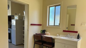 Et kompakt køkken med mikroovn, komfur og køleskab. Et lille spiseområde med stol og bord ligger ved siden af køkkenet. Et vindue med matteret glas og et spejl over vasken er synligt.