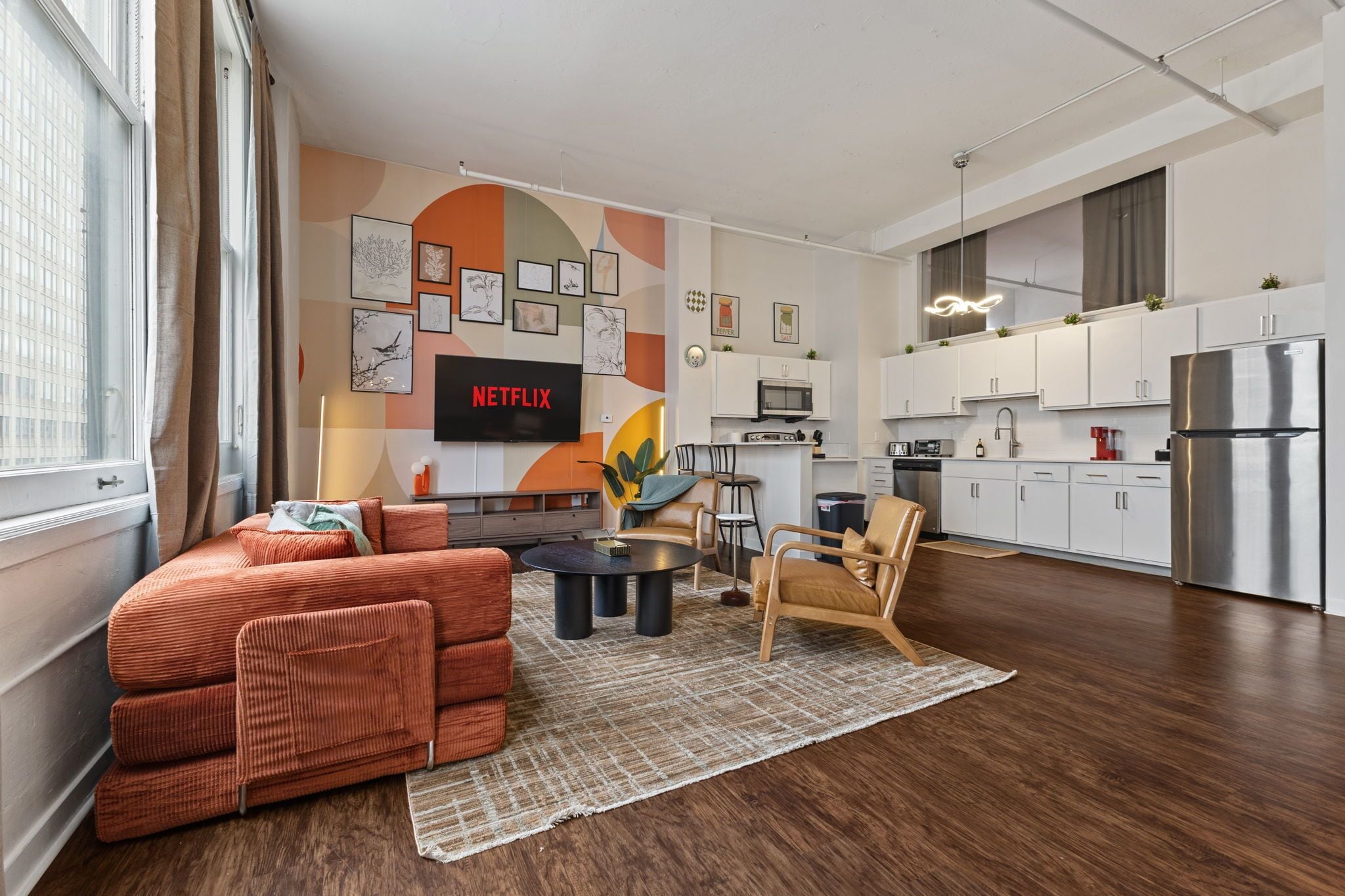 A modern living room with a sofa, coffee table, and chairs, a kitchen with white cabinets and appliances, and a wall-mounted TV displaying "NETFLIX".