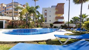 Una piscina con sillones de descanso, palmeras y un edificio con balcones.