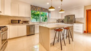 Una cocina moderna con gabinetes blancos, una isla central con taburetes, electrodomésticos de acero inoxidable e iluminación colgante.