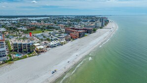 Vue aérienne d’une ville côtière avec une vaste plage de sable, un port de plaisance et divers bâtiments, notamment des hôtels et des zones résidentielles.