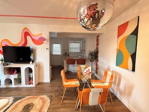 Un soggiorno con televisore, poltrone arancioni, un tavolo in vetro, una sfera da discoteca e una cucina sullo sfondo.