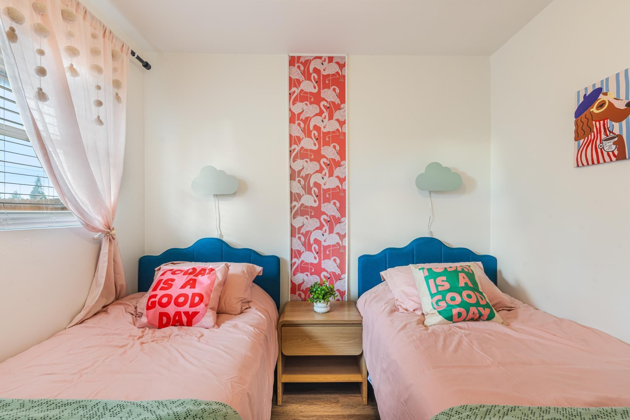 A bedroom with two beds, each with a pillow saying "Today is a good day," a small table with a plant, and a wall with a flamingo pattern.
