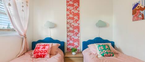 A bedroom with two beds, each with a pillow saying "Today is a good day," a small table with a plant, and a wall with a flamingo pattern.