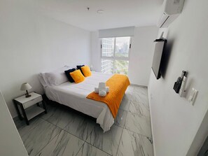 A hotel room with a bed, a nightstand, a lamp, a window with a view of buildings, and a mounted television. - Balboa Avenue Sky Suites (Panama City)