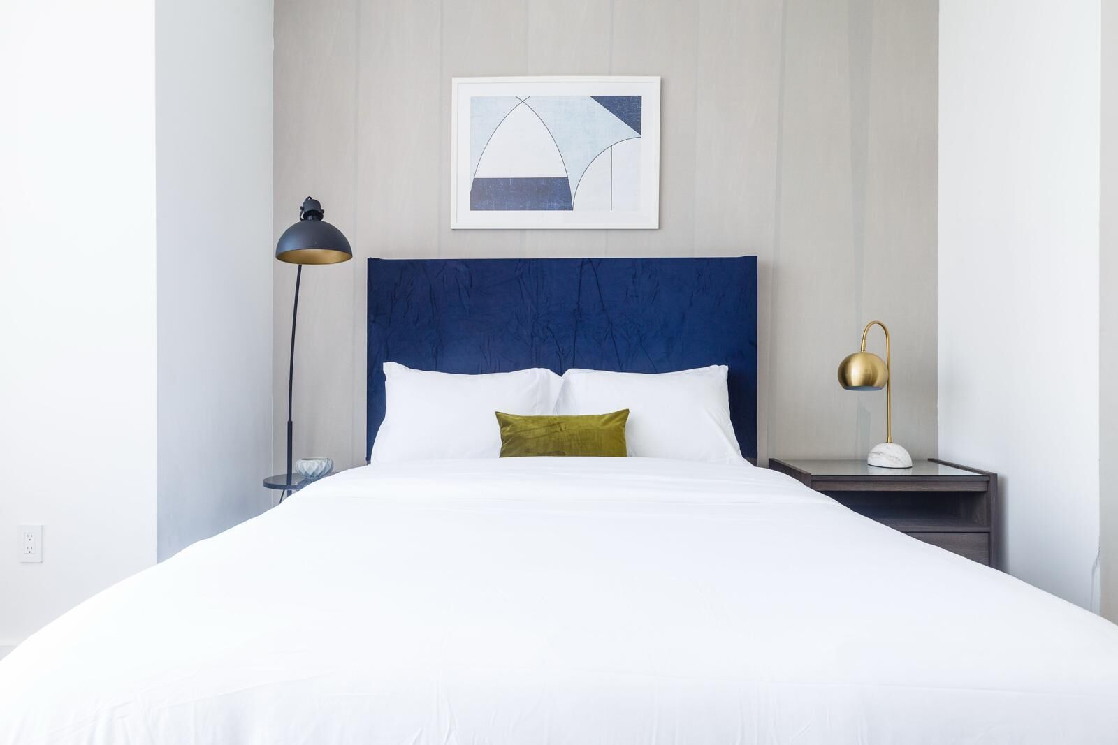 A neatly made bed with white linens, a blue headboard, and a single green pillow.