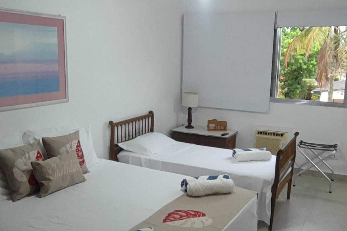A hotel room with two beds, a nightstand, a lamp, a radiator, and a window with a view of palm trees.