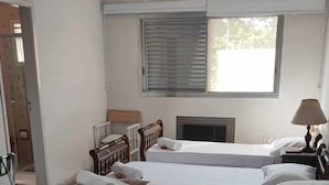 Quarto de hotel com três camas, um ventilador de teto, uma janela com persianas e um radiador.