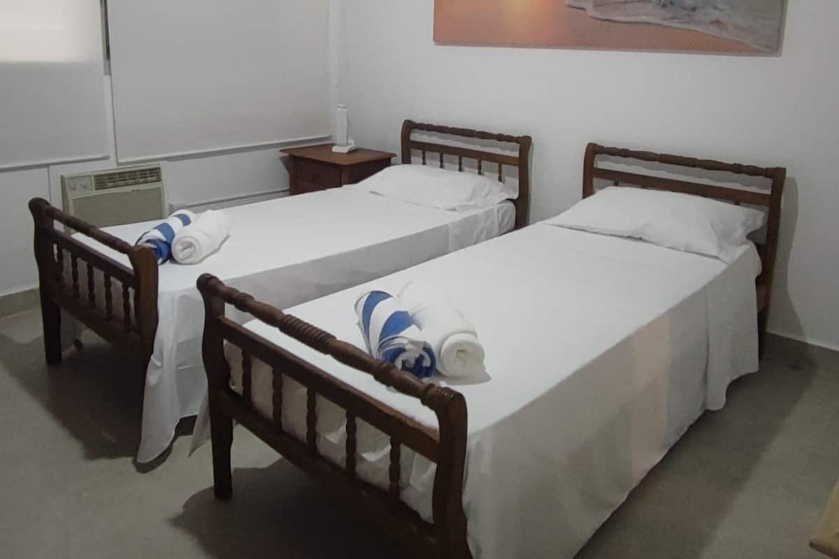 A room with two single beds, a ceiling fan, a painting of a beach scene, a window, and a radiator.