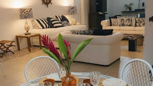 Uma área de refeições com uma mesa redonda posta para quatro pessoas, rodeada por cadeiras de vime branco. Há um vaso decorativo com flores sobre a mesa, e uma televisão é visível ao fundo.