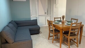 A living room with a grey sectional sofa, a wooden dining table with four chairs, and a window with blinds. - Green House Sea (Bisceglie)