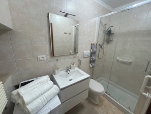 Un bagno moderno con lavandino, specchio e WC bianchi. Include una cabina doccia in vetro e un portasciugamani con asciugamani bianchi.