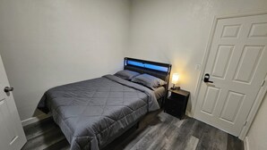 Quarto com cama, mesa de cabeceira, porta e piso de madeira.