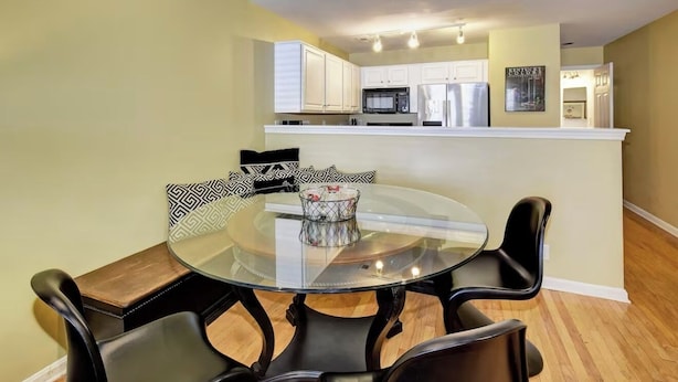 A dining area with a glass table and black chairs, a kitchen with white cabinets and stainless steel appliances, and a framed picture on the wall. - Charming Townhouse in Louisville – Explore the Urban Oasis! (Louisville)