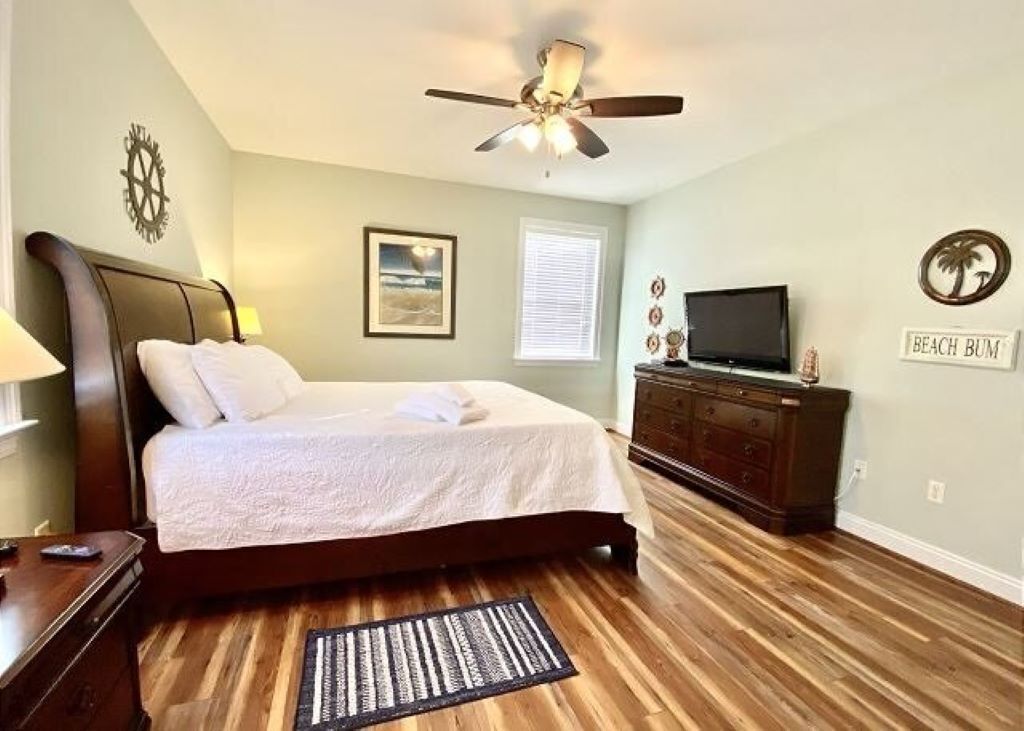 A bedroom with a bed, a ceiling fan, a dresser, a television, and a wall with a sign that reads "BEACH BUM".