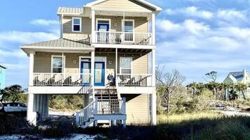 A two-story beach house with a balcony on the upper floor, a front door, and a staircase leading to the entrance.