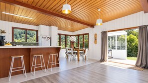 A modern kitchen with wooden beams, a bar area with stools, a dining table with chairs, and large windows offering a view of greenery. - Waterfront Bach | Deck, Kayaks & Pool Table | 15min drive from Picton (Whatamango Bay)