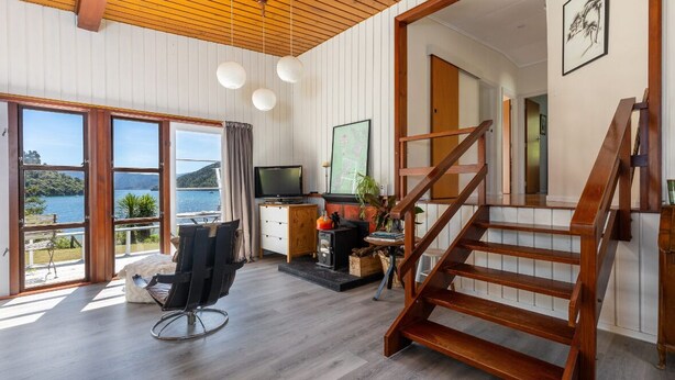 A spacious living area with a wooden staircase, large windows overlooking a lake, a television, and a cozy seating area. - Waterfront Marlborough Sounds Retreat 10min drive from Picton (Whatamango Bay)
