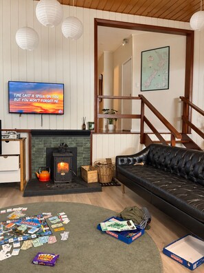 A living room with a fireplace, a sofa, a television, and a Monopoly game on the floor. - Waterfront Bach | Deck, Kayaks & Pool Table | 15min drive from Picton (Whatamango Bay)